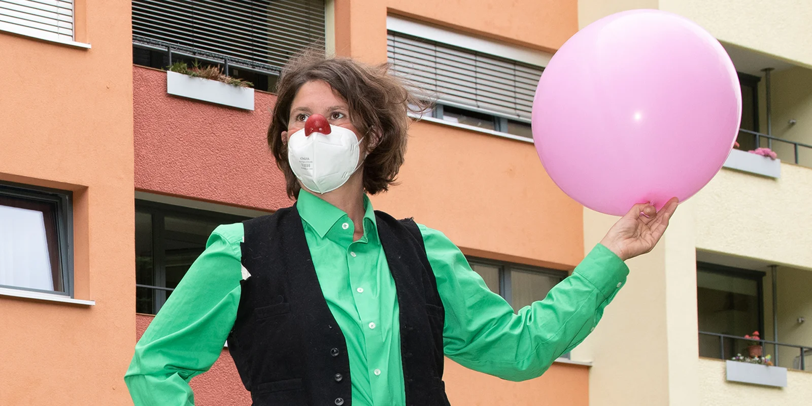 Norika Wacker trägt eine FFP2-Maske und hält einen rosa Ballon in der Hand. Sie hat eine rote Clownsnase auf ihrer FFP2-Maske.