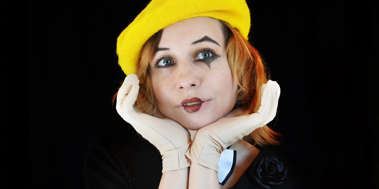 Eine junge Frau mit Clown-Schminke im Gesicht hat ihren Kopf auf ihre Hände gestützt. Sie trägt einen gelben Hut.