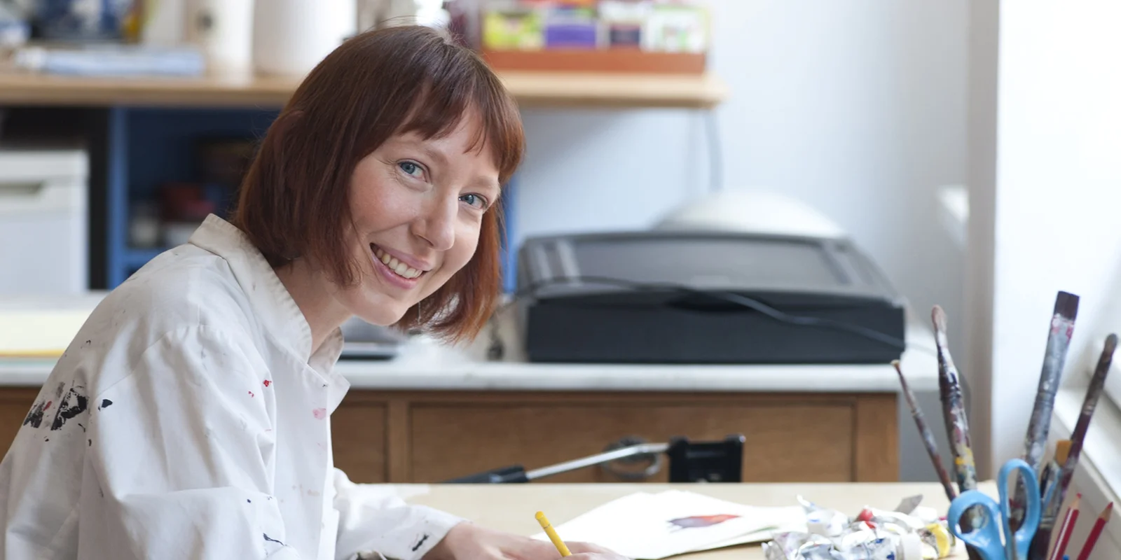 Ruth Reiche sitzt im Malerkittel zeichnend an einem Schreibtisch und lächelt in die Kamera