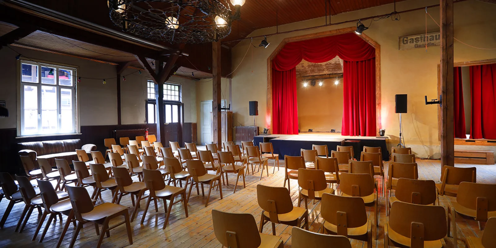 Das Studio Klawunn von Innen: eine Bühne mit rotem Vorhang, davor mehrere Reihen mit Holzstühlen. Im Hintergrund eine Empore aus dunklem Holz.