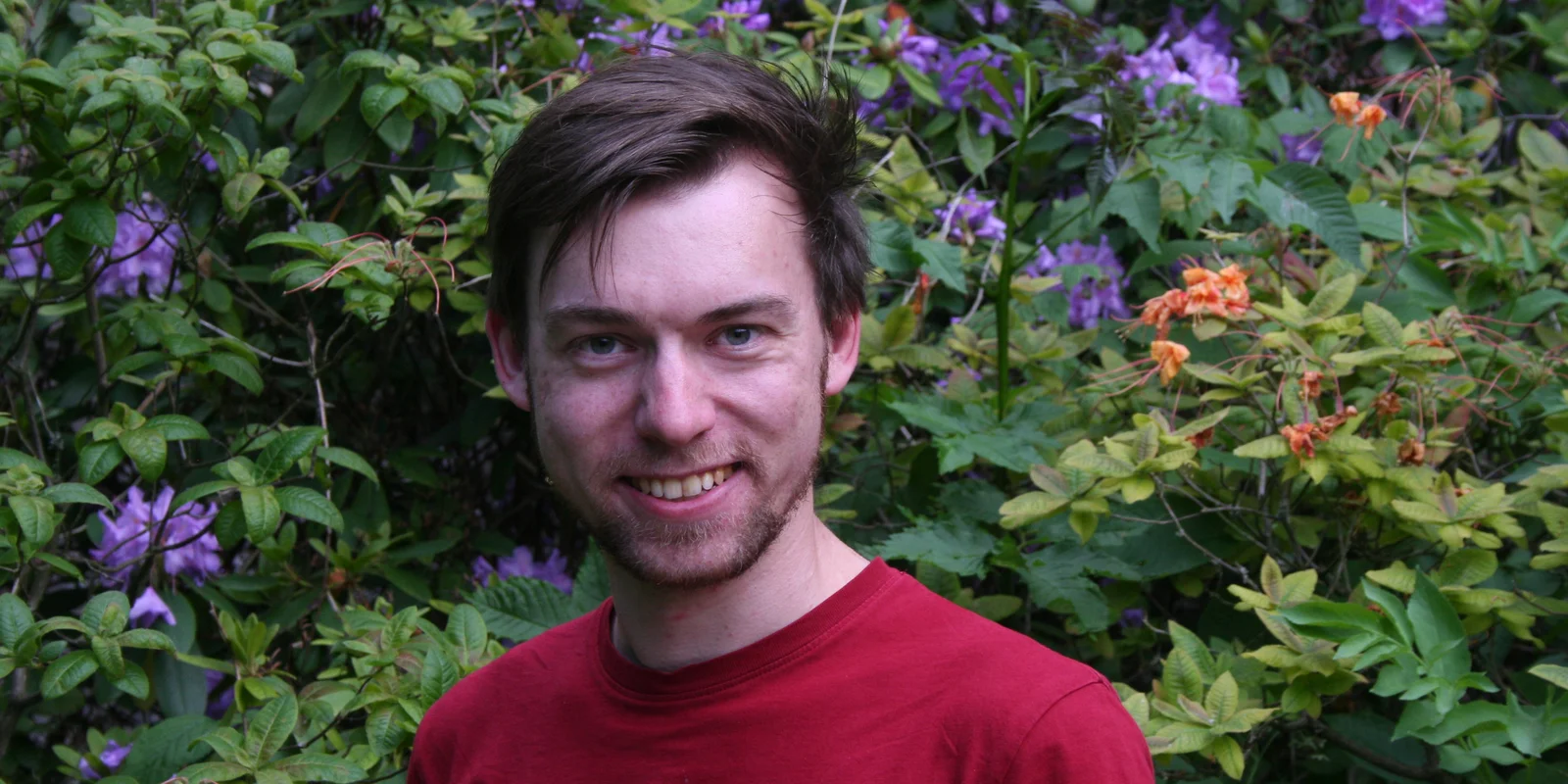 Lukas Keller lächelt mit rotem T-Shirt vor einer Blumenhecke in die Kamera